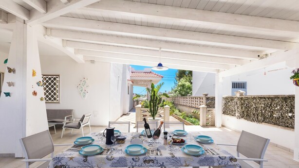 An outdoor dining area with a table set for two, surrounded by white chairs, a potted plant, and a decorative wall. - Villetta Sea Sunset by BarbarHouse (Specchiarica)