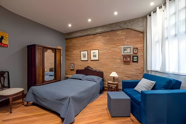 A bedroom with a bed, a blue sofa, a wooden wardrobe, and a brick wall with framed pictures. - Palacete Riachuelo (São Paulo)