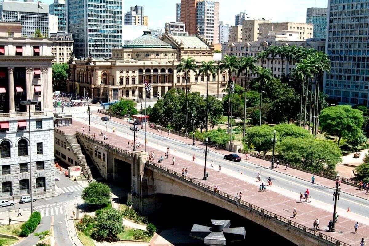Un paisaje urbano con un edificio destacado que tiene una cúpula y ventanas en arco, un puente con peatones y altas palmeras.