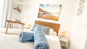 Een netjes opgemaakt bed met een opgerolde handdoek, een houten nachtkastje met een lamp, een aan de muur hangend schilderij en een kapstok.