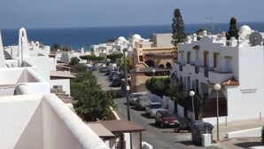 A coastal street with white buildings, parked cars, and a clear view of the sea.