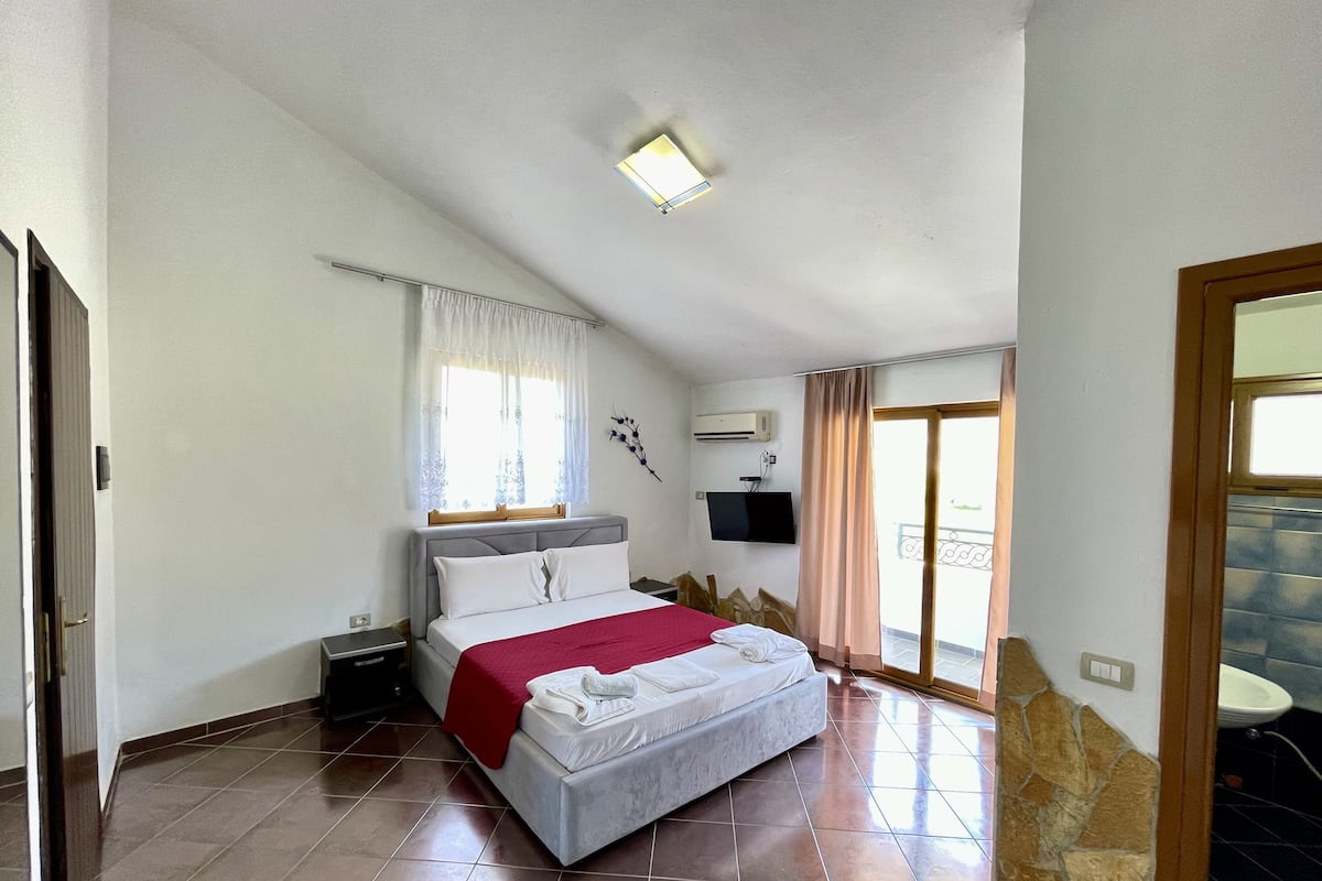A bedroom with a bed, a window with curtains, a TV mounted on the wall, and a bathroom visible through an open door.