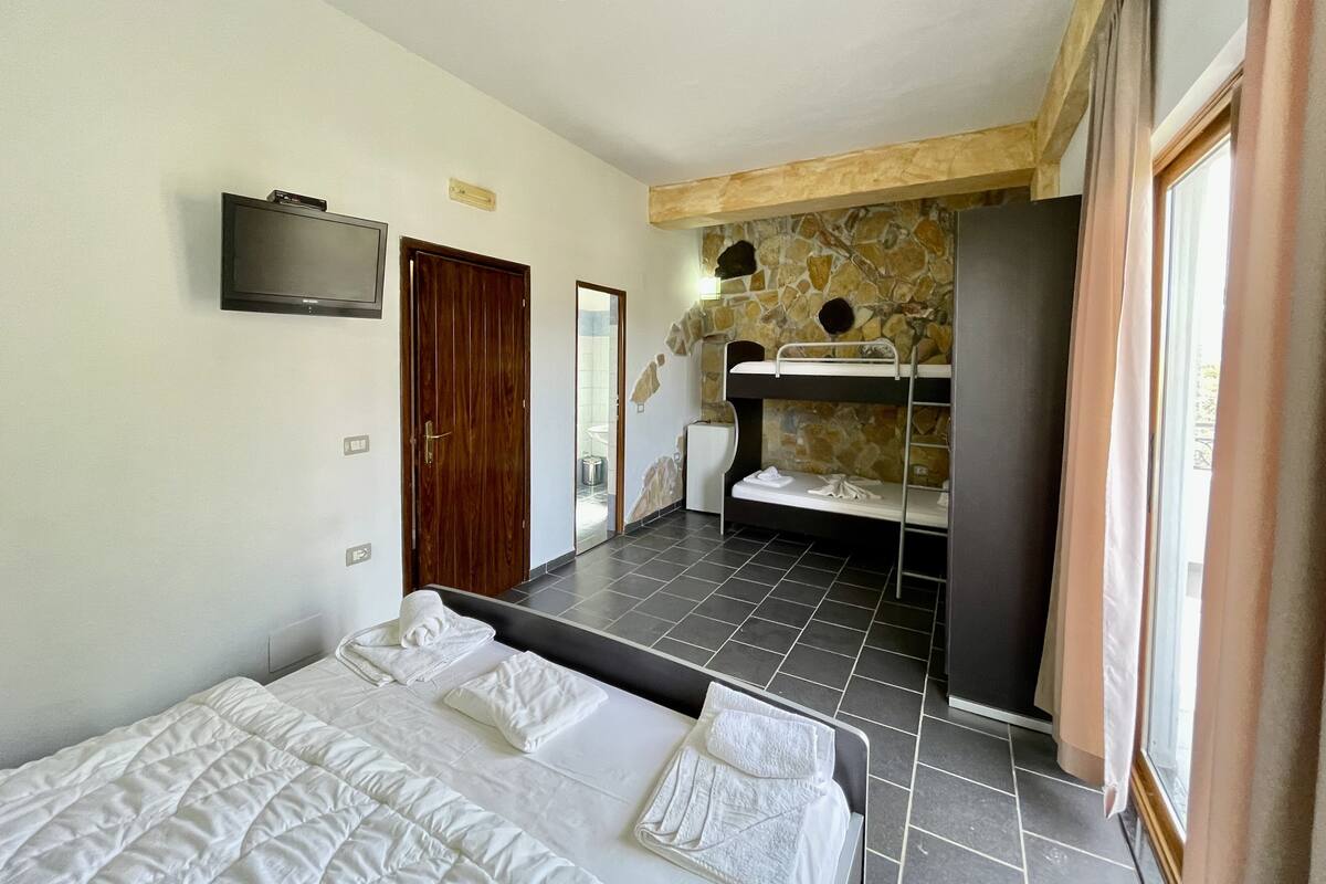 A bedroom with a bed, a television, a wooden door, a stone wall, and a bathroom visible through an open door.