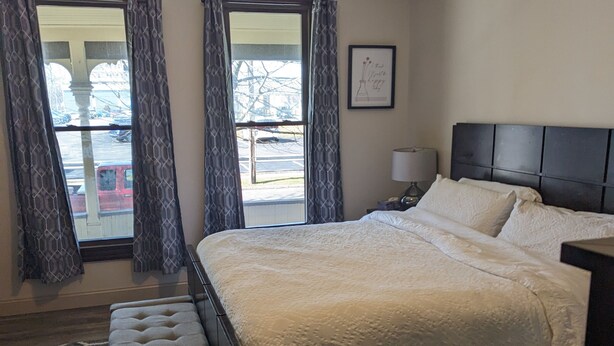 A bedroom with a large bed, a nightstand, a framed picture on the wall, and a view through the window. - Hidden Gem in Cortland (Cortland)