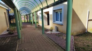 Une allée couverte avec un sol carrelé, des piliers verts et un bâtiment doté de fenêtres et d’une porte.