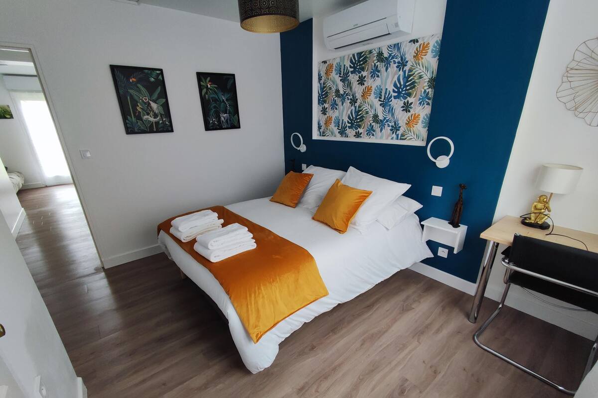 Een moderne slaapkamer met een bed, beddengoed in wit en oranje, een blauwe accentmuur en een klein bureau met een lamp.