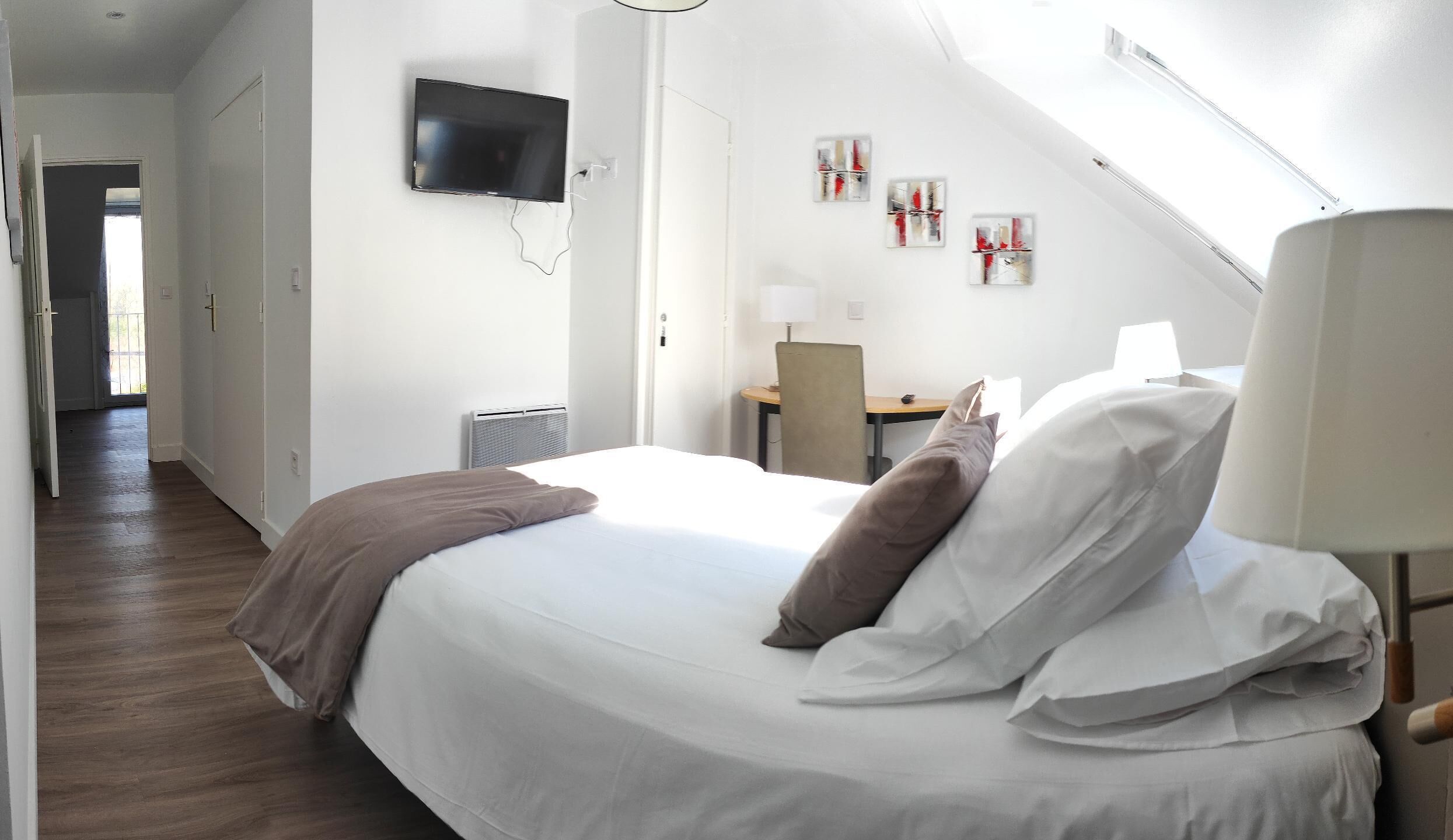 Habitación de hotel con cama, escritorio, silla, televisor montado en la pared y cuadros decorativos.