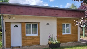 Un bâtiment à deux étages, avec un toit rouge, un revêtement en bois et une porte blanche. On y trouve un petit jardin avec des fleurs violettes et un pot de fleurs noir.
