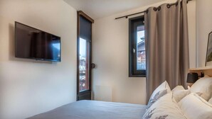 A bedroom with a bed, a flat-screen TV mounted on the wall, a window with curtains, and a view of buildings outside. - 190m from ski slopes, 1st floor, balcony, tv, ski locker, 39m², Morzine (Morzine)