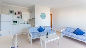 A compact living space with a kitchenette, two sofas with blue cushions, a dining table, and a wall clock.