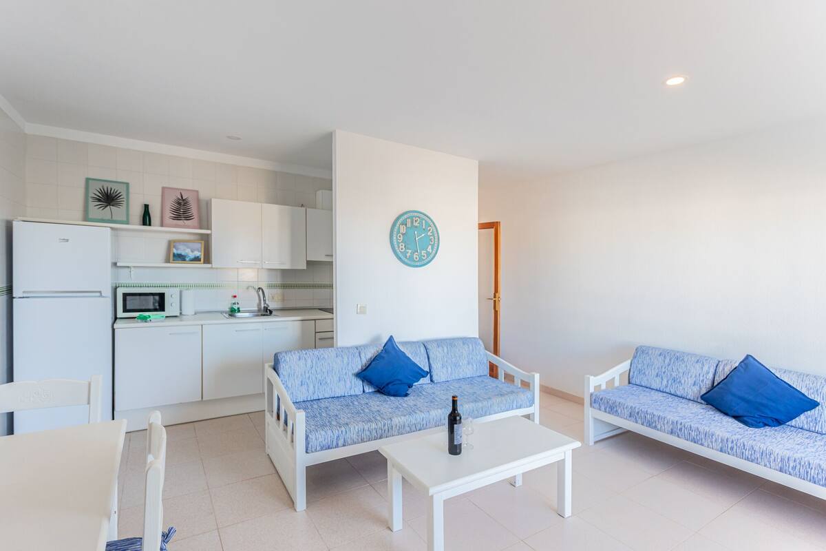 Um espaço compacto com cozinha compacta, dois sofás com almofadas azuis, uma mesa de jantar e um relógio de parede.