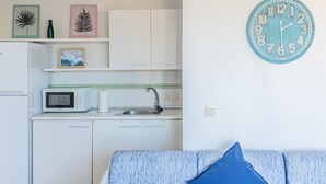 A kitchen with white cabinets, a microwave, and a sink. There is a teal clock on the wall and a blue couch with a blue pillow.