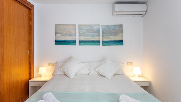 A bedroom with a bed, two bedside lamps, a wall-mounted air conditioner, and three paintings of the sea.