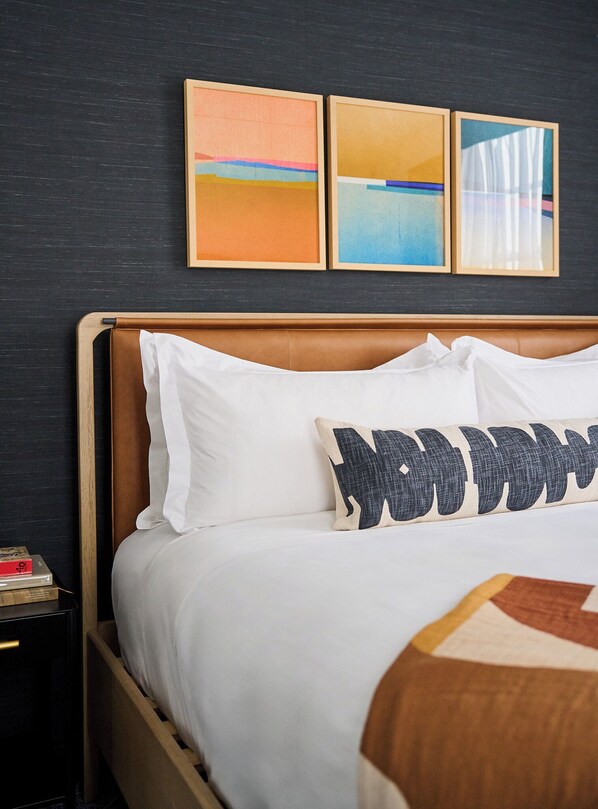 A neatly made bed with white linens and a pillow with a bold design, a wooden headboard, and three framed artworks on the wall above.