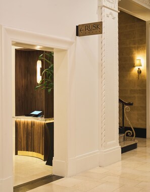 A reception desk with a wooden finish and a digital screen, a sign indicating 'RUSK HOUSE', and a staircase with a decorative metal railing.