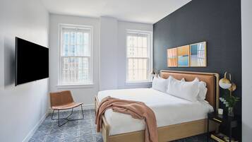 A modern hotel room with a large bed, two windows, a flat-screen TV, a chair, and a bedside table with a lamp.