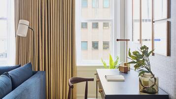 A modern, compact living space with a sofa, a dining table, and a chair. There is a window with curtains, a lamp, and a plant on the table.