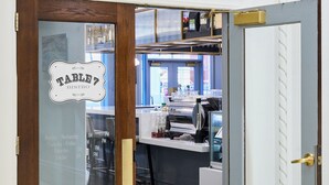 En liten restaurang med en trädörr och ett glasfönster. Inuti finns en bardisk med en kaffemaskin och diverse andra saker.