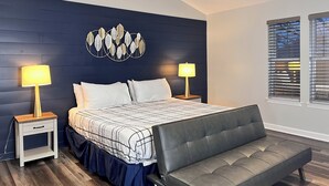 Een slaapkamer met een donkerblauwe accentmuur, een bed met een zwart-wit beddeken, twee bedlampen, een bankje en een raam met jaloezieën.