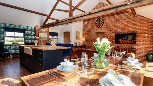 A dining area with a wooden table set for a meal, a brick wall, and a large window. - Beth's Cottage (Malton)