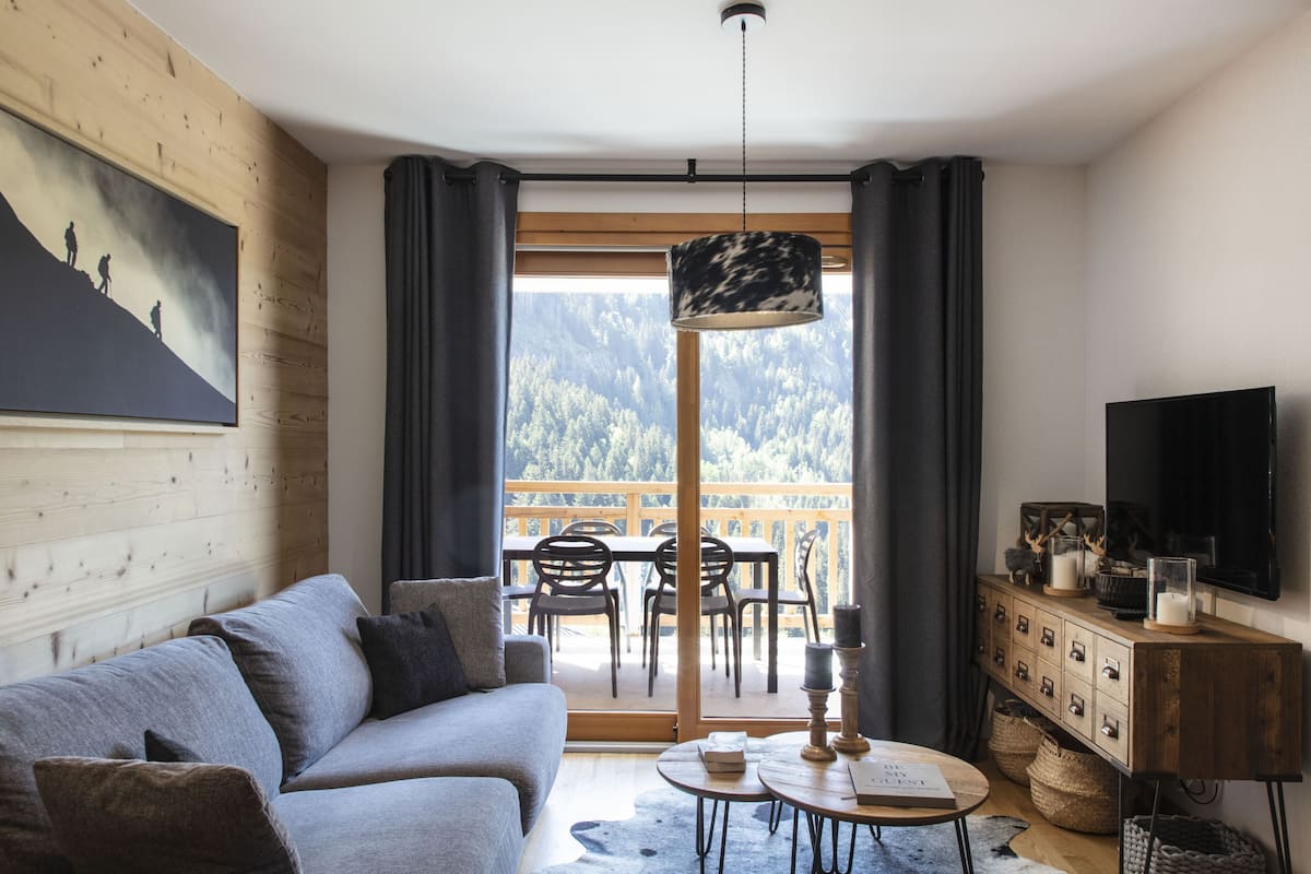 A modern living room with a sofa, a dining table, and chairs, a television, and a view of a forested mountain through a large window.