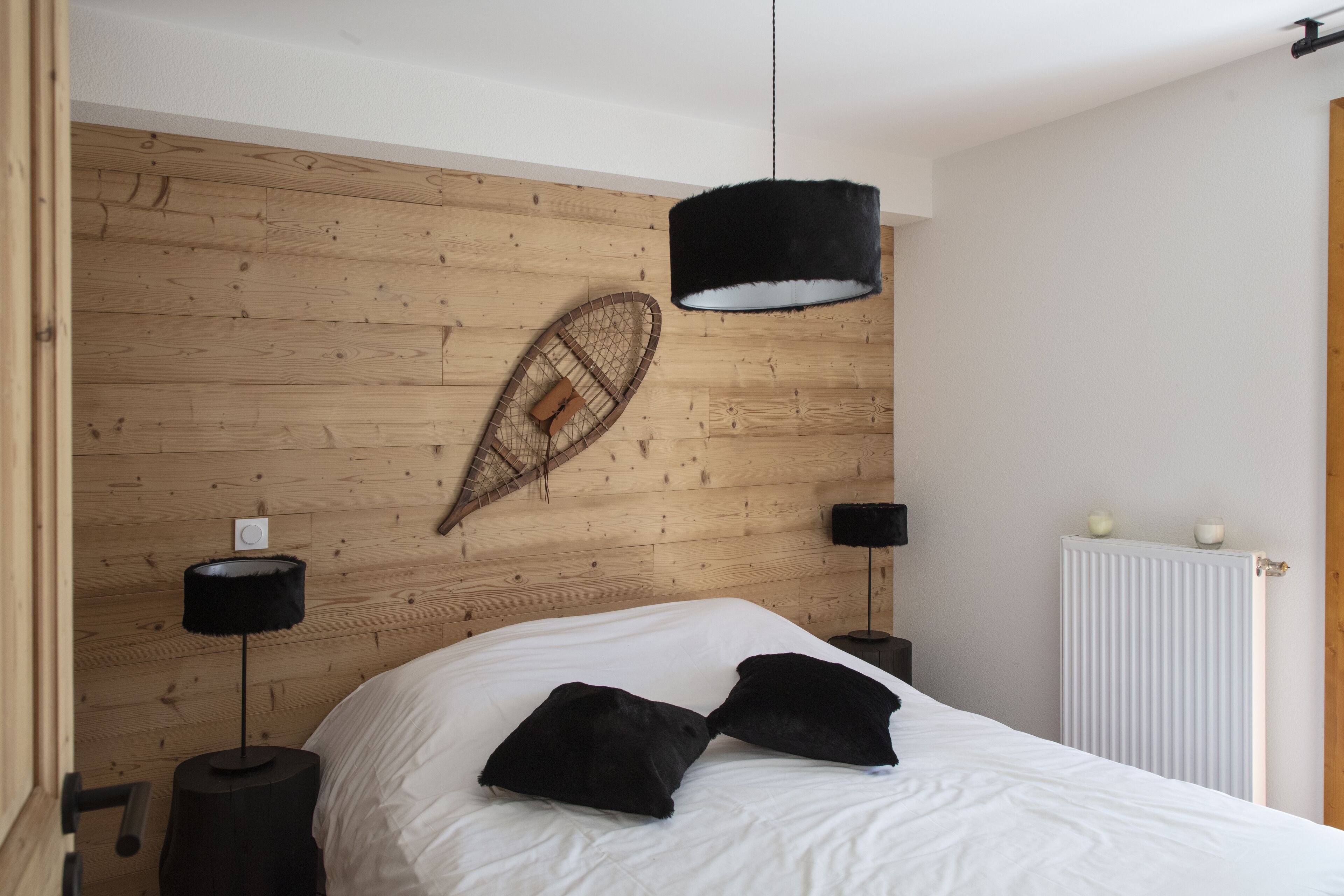 Une chambre avec une tête de lit en bois, un lit avec une literie blanche, deux oreillers noirs, une lampe noire suspendue et un luminaire fixé au mur.