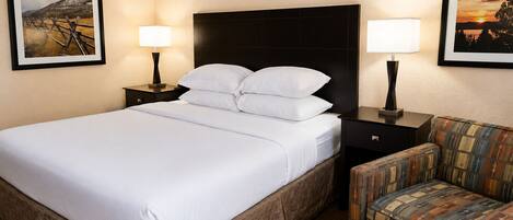 A hotel room with a large bed, two bedside tables with lamps, a patterned armchair, and two framed pictures on the walls.