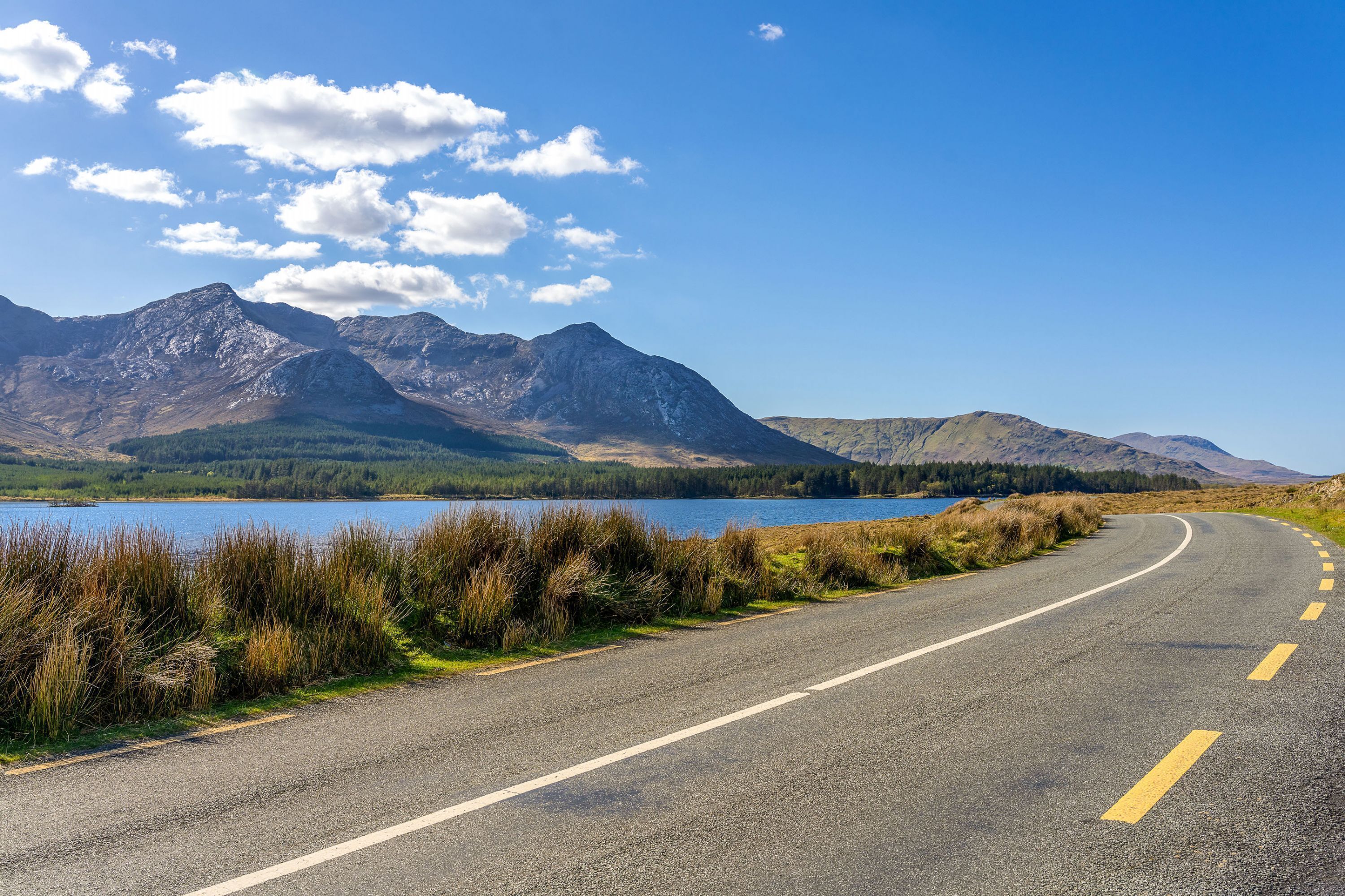 Une route sinueuse, avec des montagnes en arrière-plan et une étendue d’eau sur la gauche.