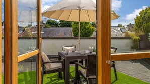 Een terras met een tafel, stoelen, een parasol en een glazen deur die naar een tuin leidt.