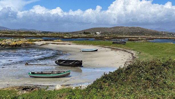 Un paysage côtier avec des bateaux sur une plage de sable, des zones herbeuses et des collines lointaines sous un ciel partiellement nuageux.