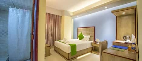 Quarto de hotel com cama, escrivaninha, cadeira, espelho e área de chuveiro.