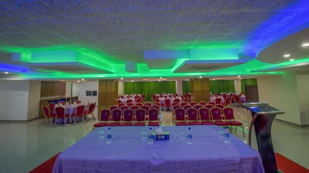 A banquet hall with rows of red chairs, a long table with water bottles, and a stage with a podium.