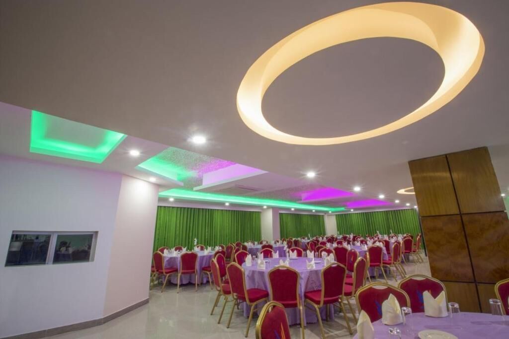A banquet hall with round tables and red chairs, green curtains, and a modern ceiling design.