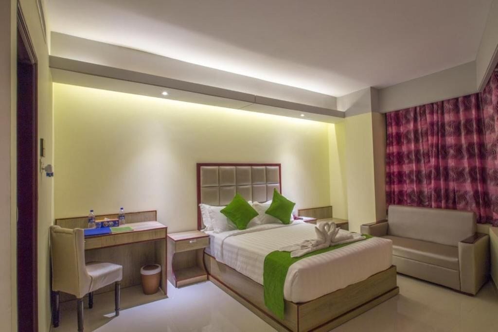 A hotel room with a bed, a desk, a chair, a sofa, and a trash bin.