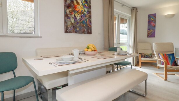 Uma sala de jantar moderna com uma mesa branca, cadeiras verde-água e um banco. Há uma tigela de frutas sobre a mesa, e obras de arte nas paredes.
