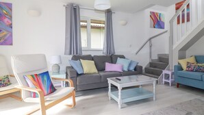 Uma sala de estar moderna com um sofá cinza, almofadas coloridas, uma poltrona branca, uma mesa de centro de vidro e uma escada com corrimão branco.