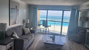 Un salon avec un canapé, une table basse et un fauteuil. Une grande fenêtre offre une vue sur la plage et l’océan.