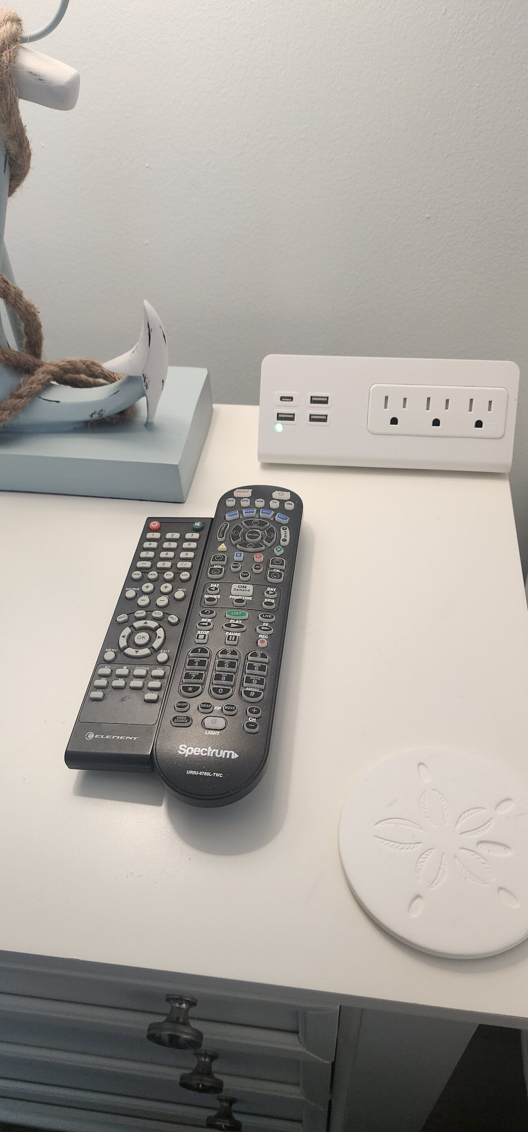 Et hvidt bord med to sorte Spectrum fjernbetjeninger, en hvid stikdåse og en hvid dekorativ tallerken.