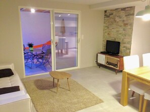 Sala de estar com sofá, uma mesinha, televisão sobre um m óvel, e porta de vidro deslizante que dá para uma varanda com móveis de exterior.