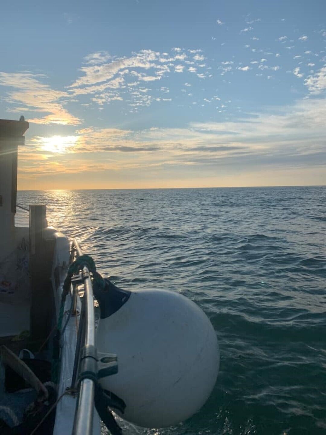 A view of the sea from a ship with a white buoy in the foreground and a sunset in the background.