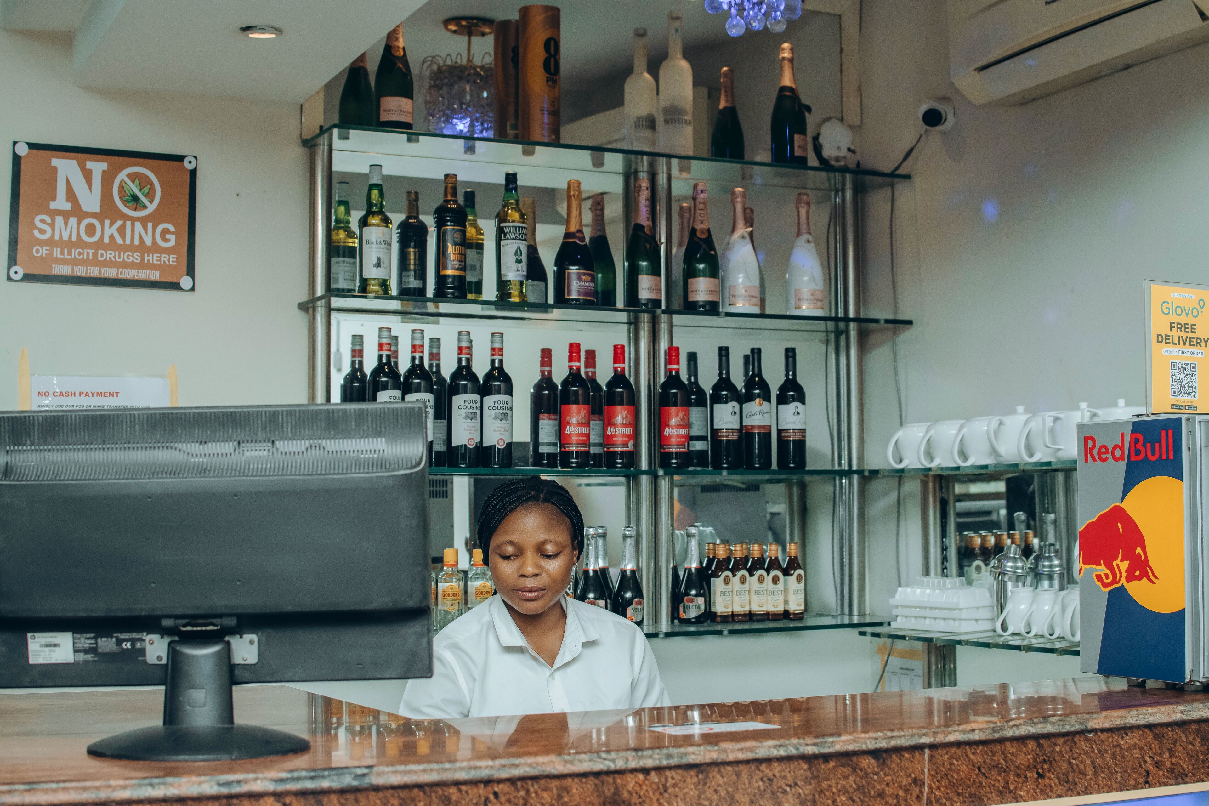 Un espace bar avec une variété de bouteilles d’alcool exposées sur des étagères, un panneau indiquant qu’il est interdit de fumer et un réfrigérateur Red Bull.