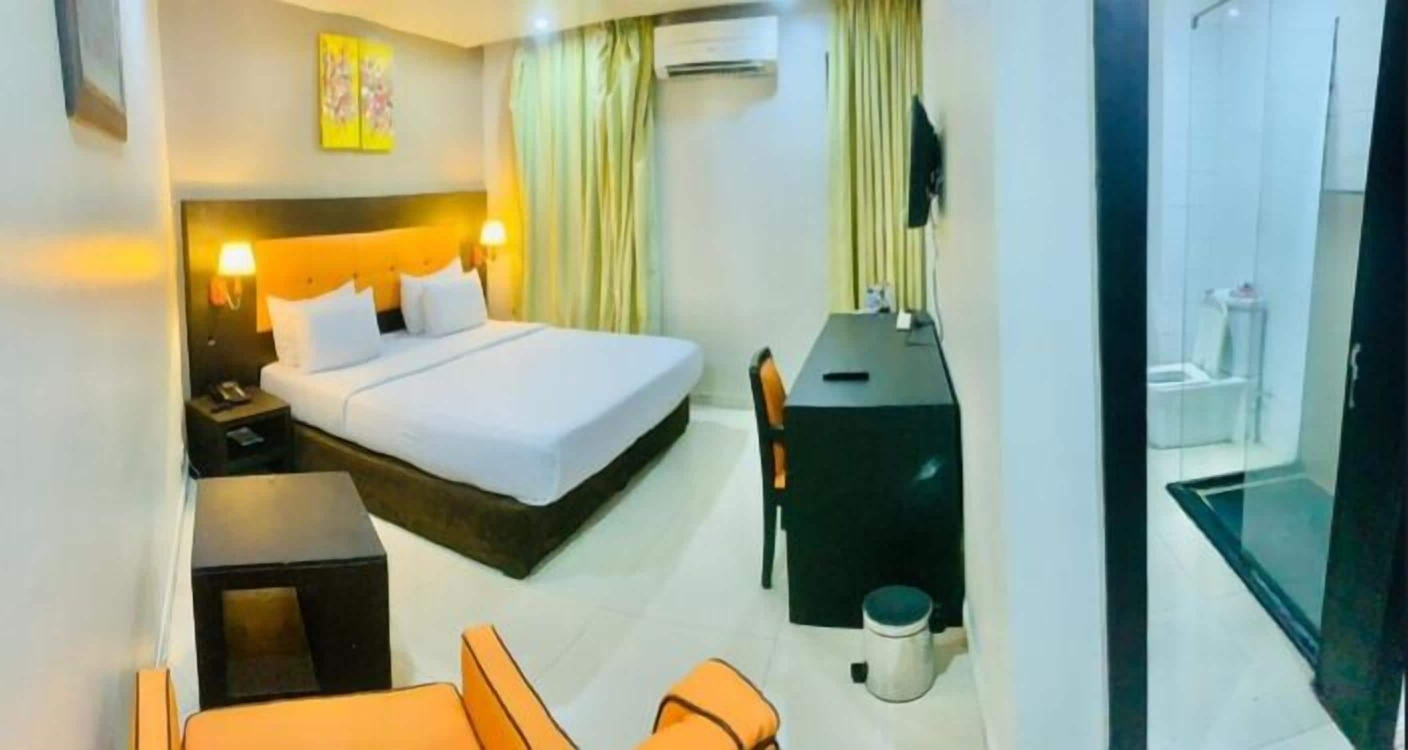 A hotel room with a bed, a desk, a chair, a small table, a trash bin, and a shower area.