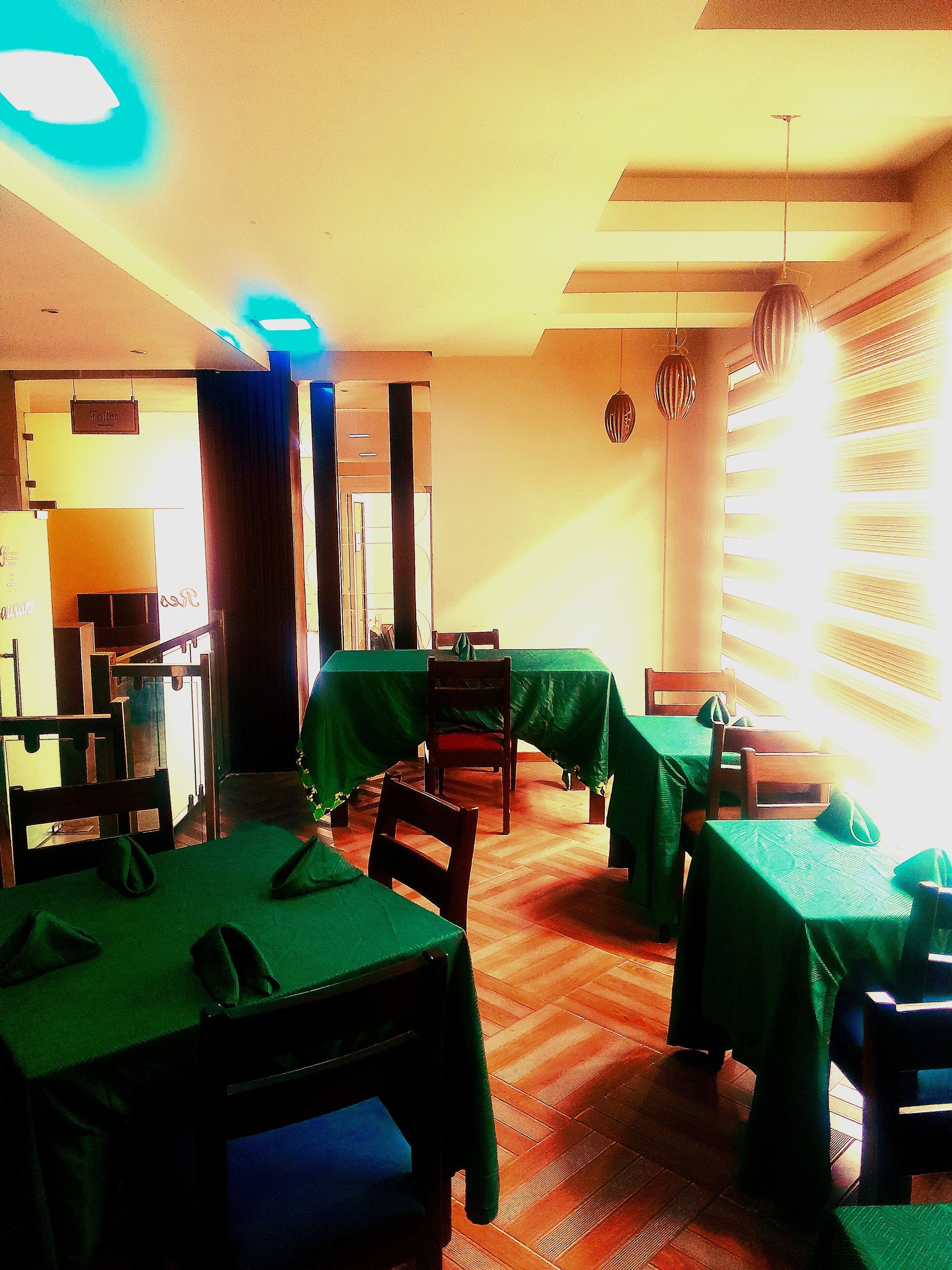 A dining area with tables covered in green tablecloths, wooden chairs, and hanging pendant lights.