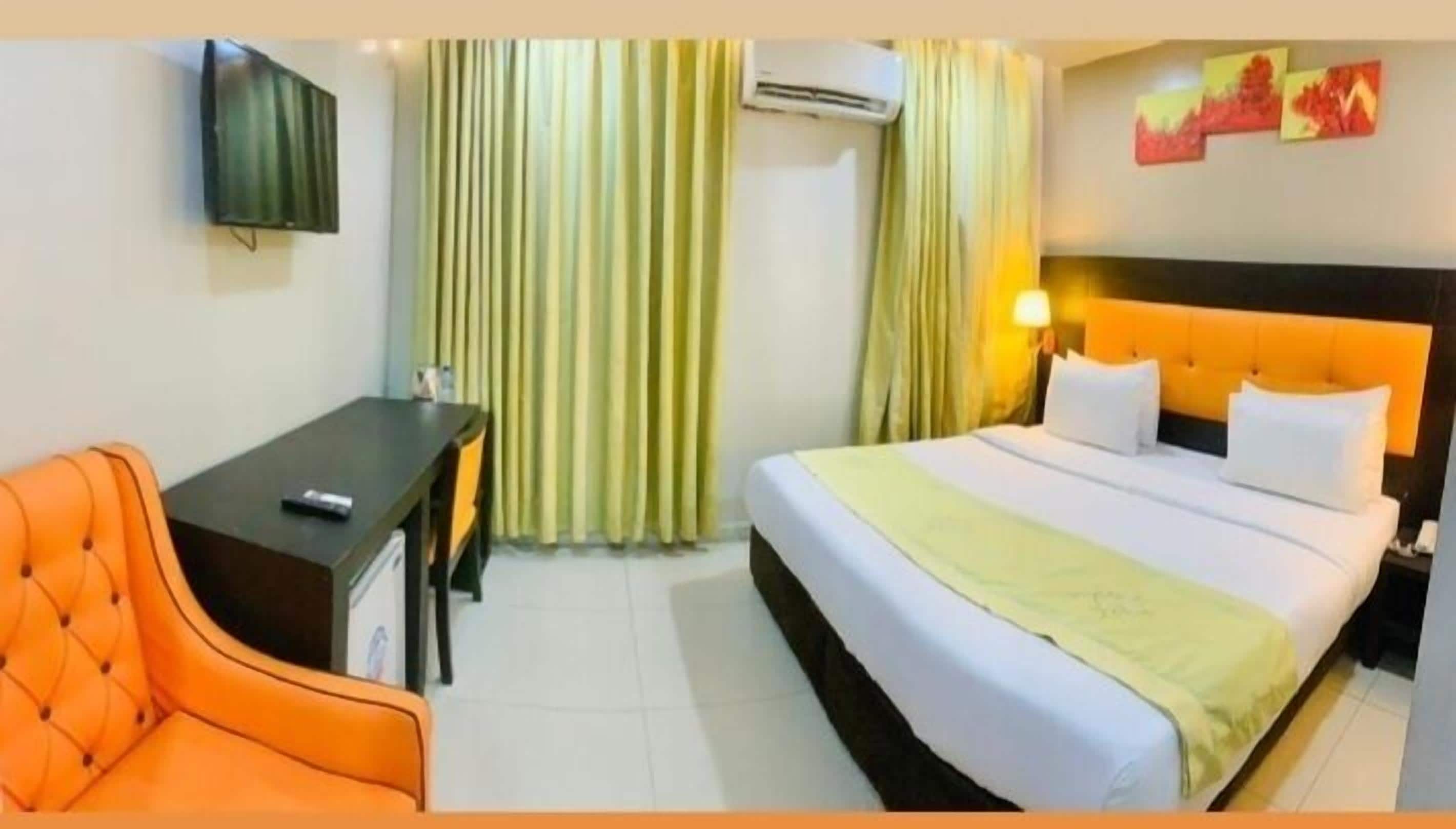 A hotel room with a bed, a desk, an orange chair, curtains, and artwork on the wall.