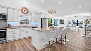 A modern kitchen with white cabinetry, a central island with bar stools, a large clock, and a dining area with a table and chairs. - Seaside Sanctuary at Jupiter Inlet / Dubois Park (Jupiter)