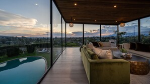 Sala de estar moderna com grande parede de vidro, piscina e vista para a paisagem urbana.