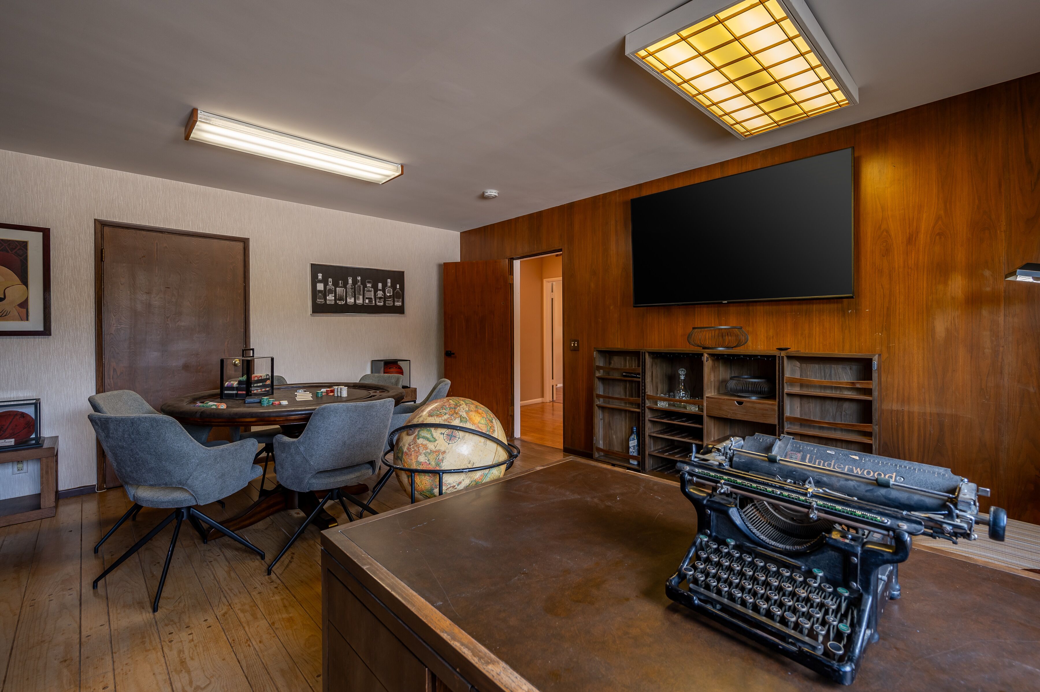 Une pièce avec un bureau en bois, un globe terrestre, une machine à écrire ancienne et un téléviseur à écran plat fixé au mur.