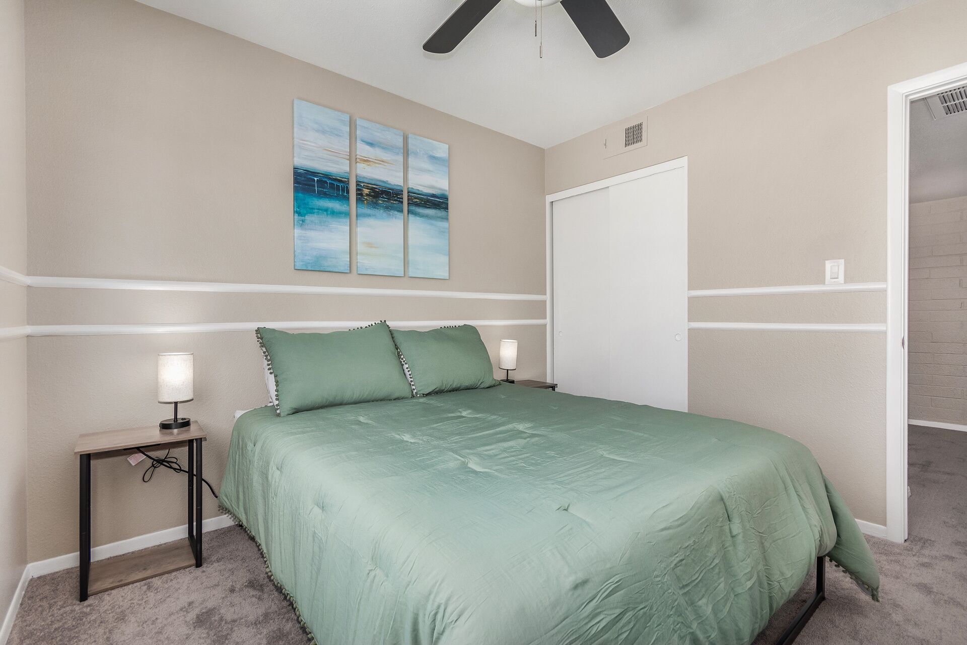 A bedroom with a bed, nightstands, a ceiling fan, a door, and a wall with three paintings.