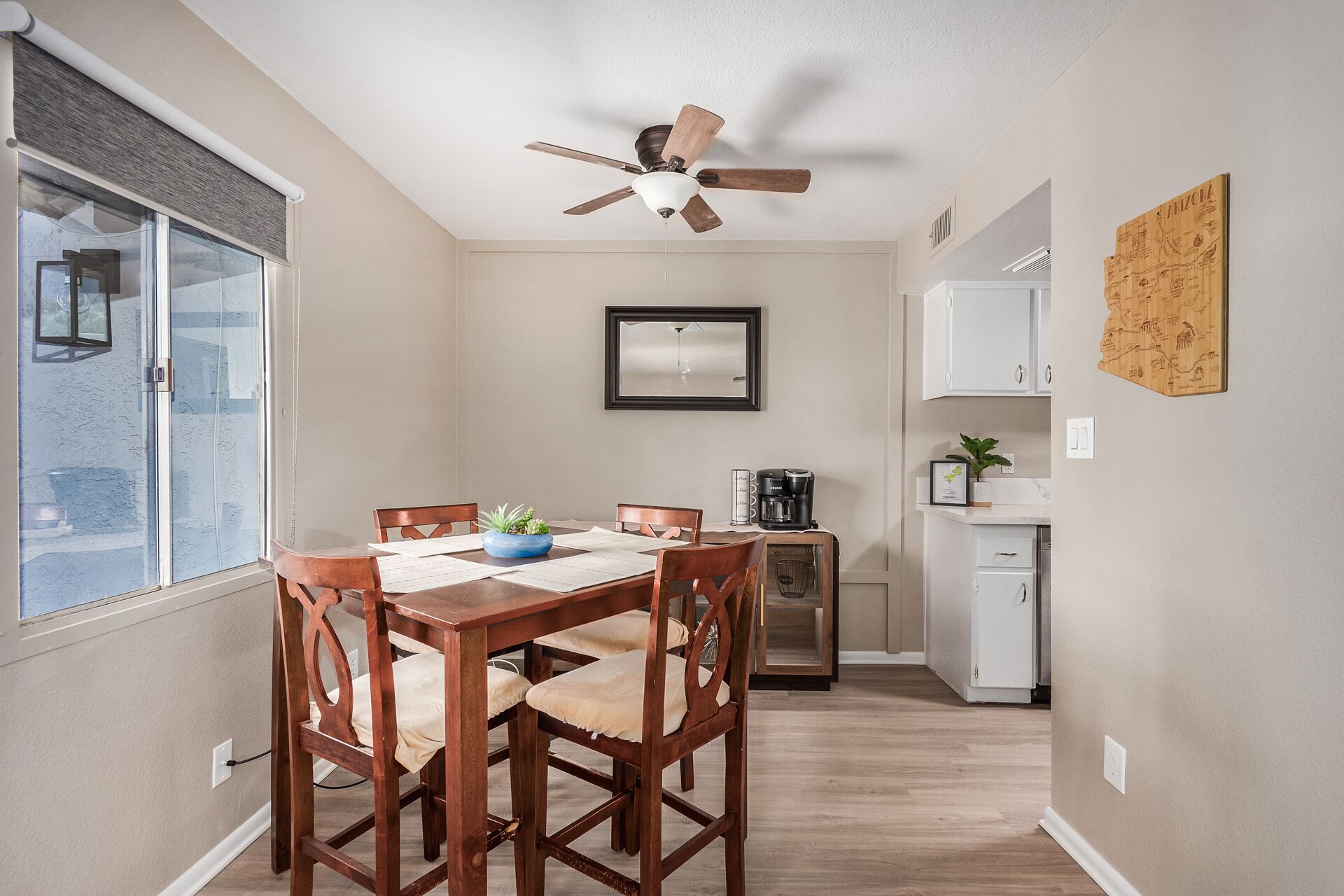 A dining area with a wooden table and chairs, a ceiling fan, a window with blinds, and a kitchen area with cabinets and a countertop.