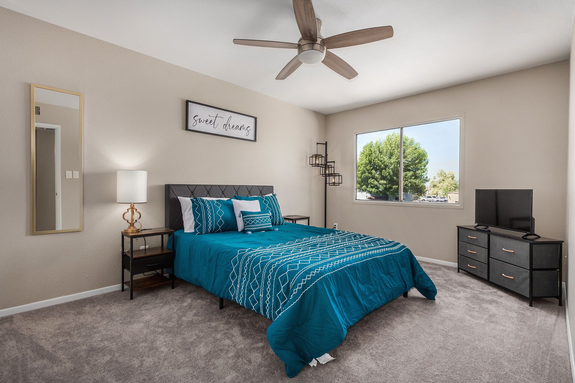 A bedroom with a bed, a ceiling fan, a window with a view, a mirror, a bedside table with a lamp, and a dresser.